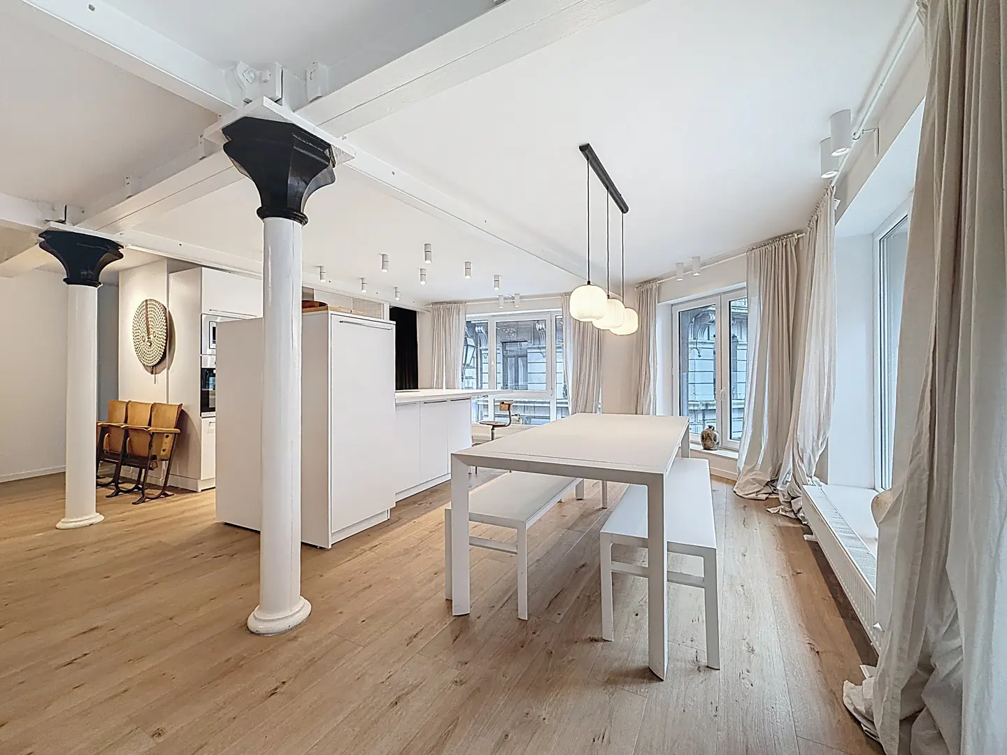 Bright, modern loft with white walls, wood floors, and large windows. A white table and benches sit under pendant lights. Columns add architectural detail.