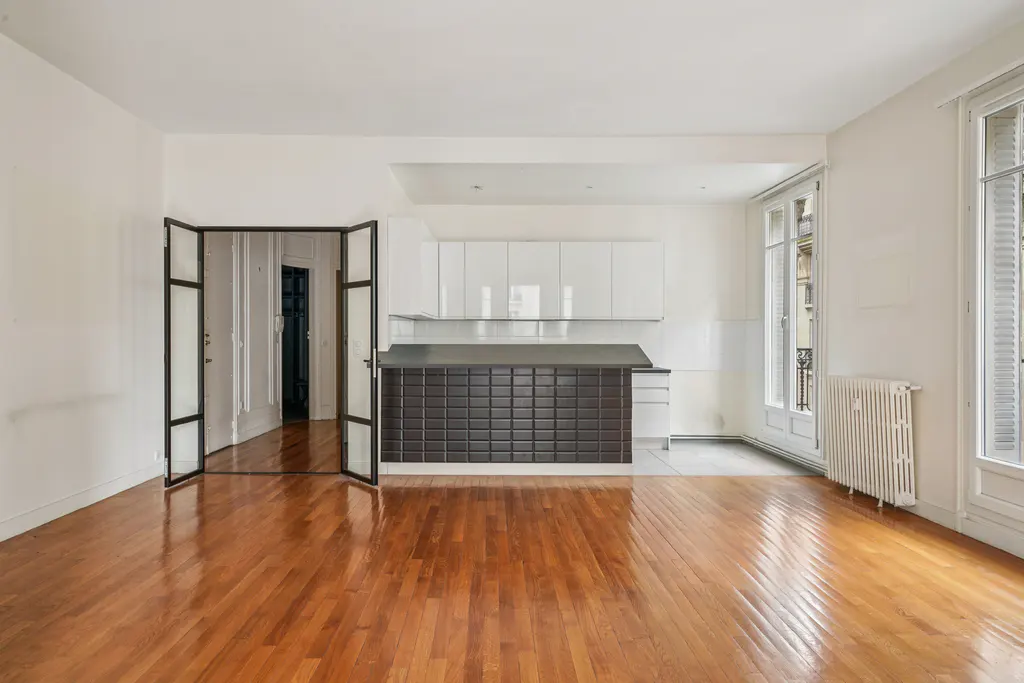 Bright, empty room with hardwood floors, white walls, and a modern kitchen. Black-framed glass doors and windows let in natural light.