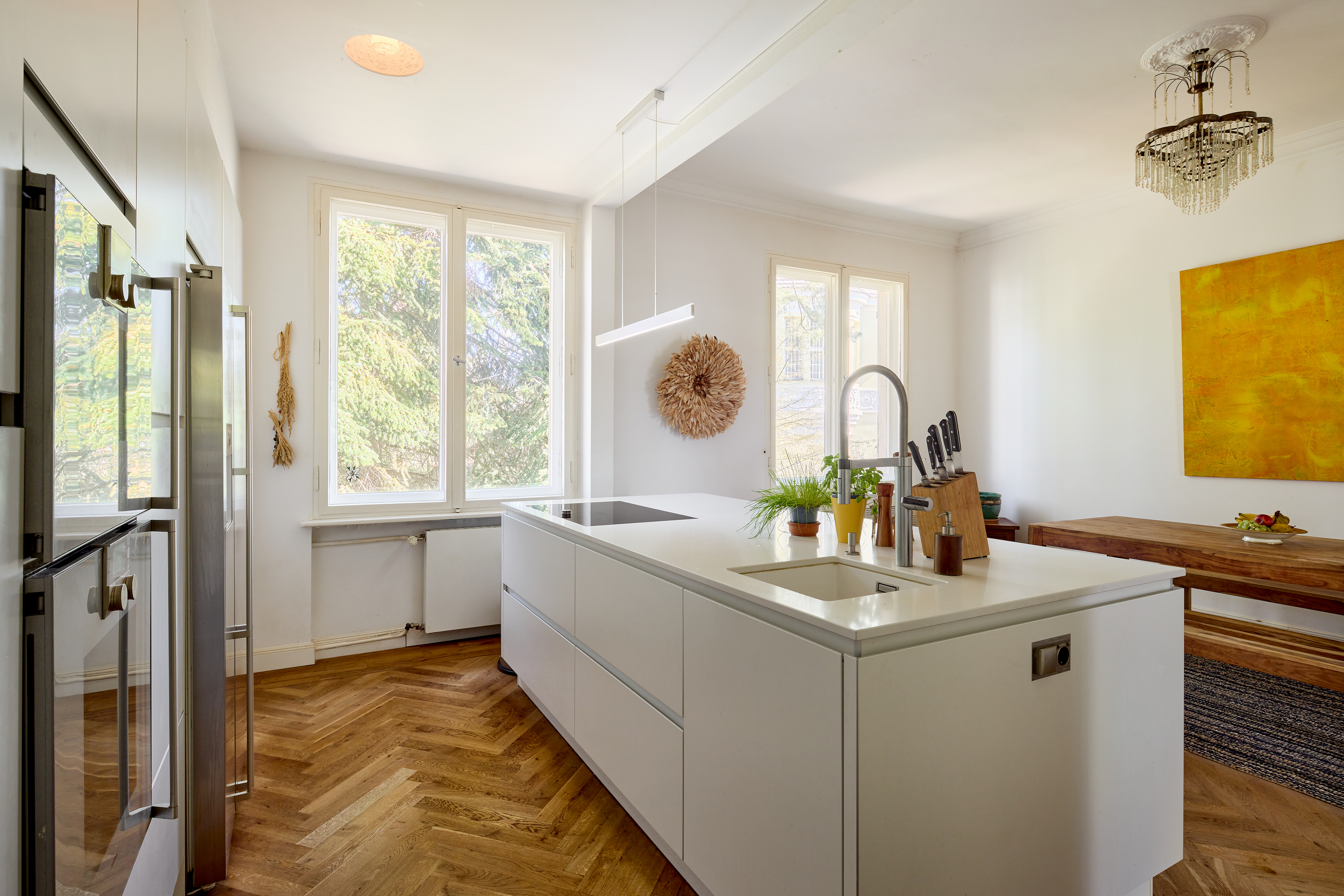 Modern white kitchen with a central island, stainless steel appliances, herringbone wood floors, and a bright yellow painting.