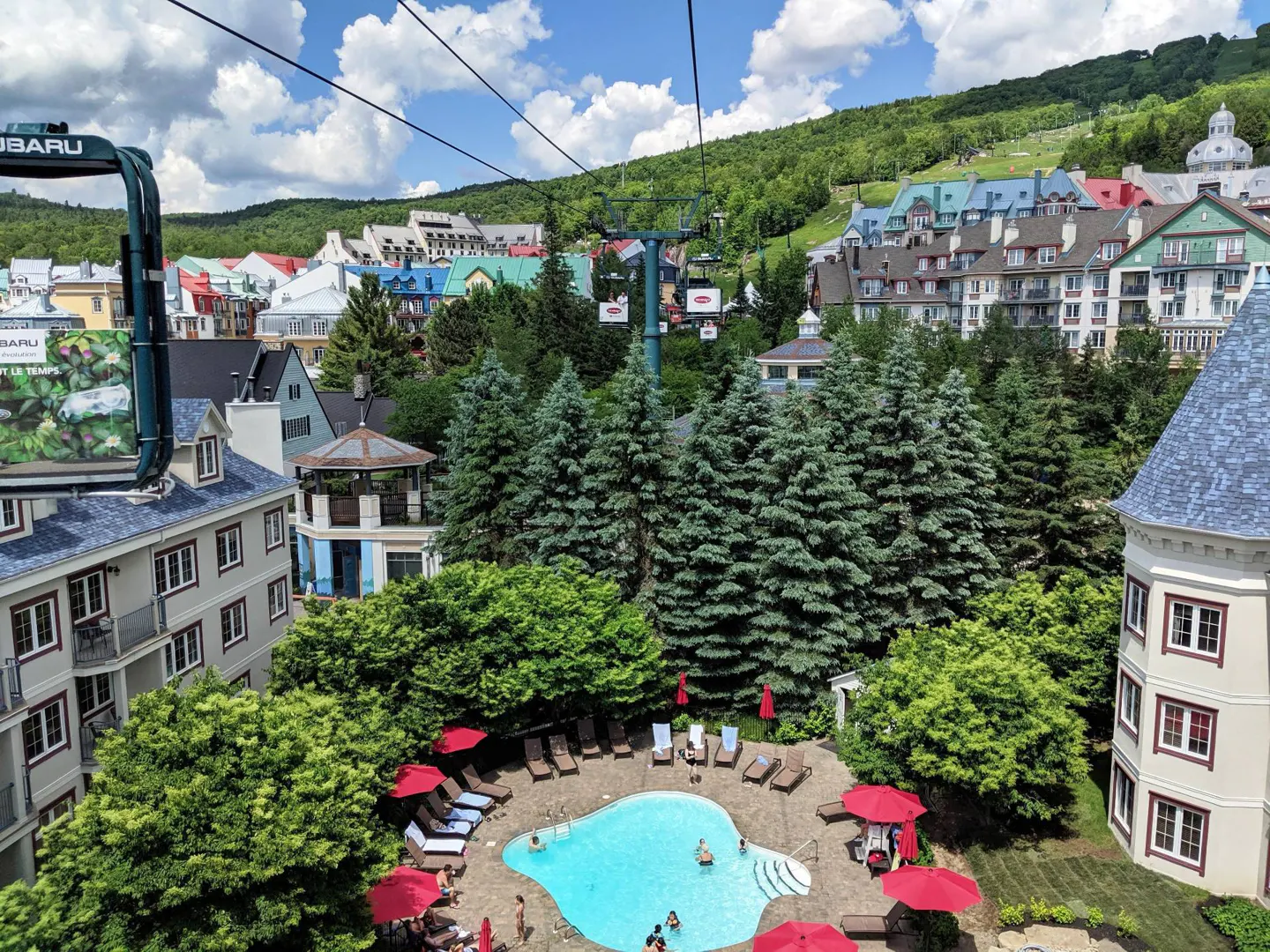 View from a ski lift of a resort town with a pool, lounge chairs, and colorful buildings on a sunny day.