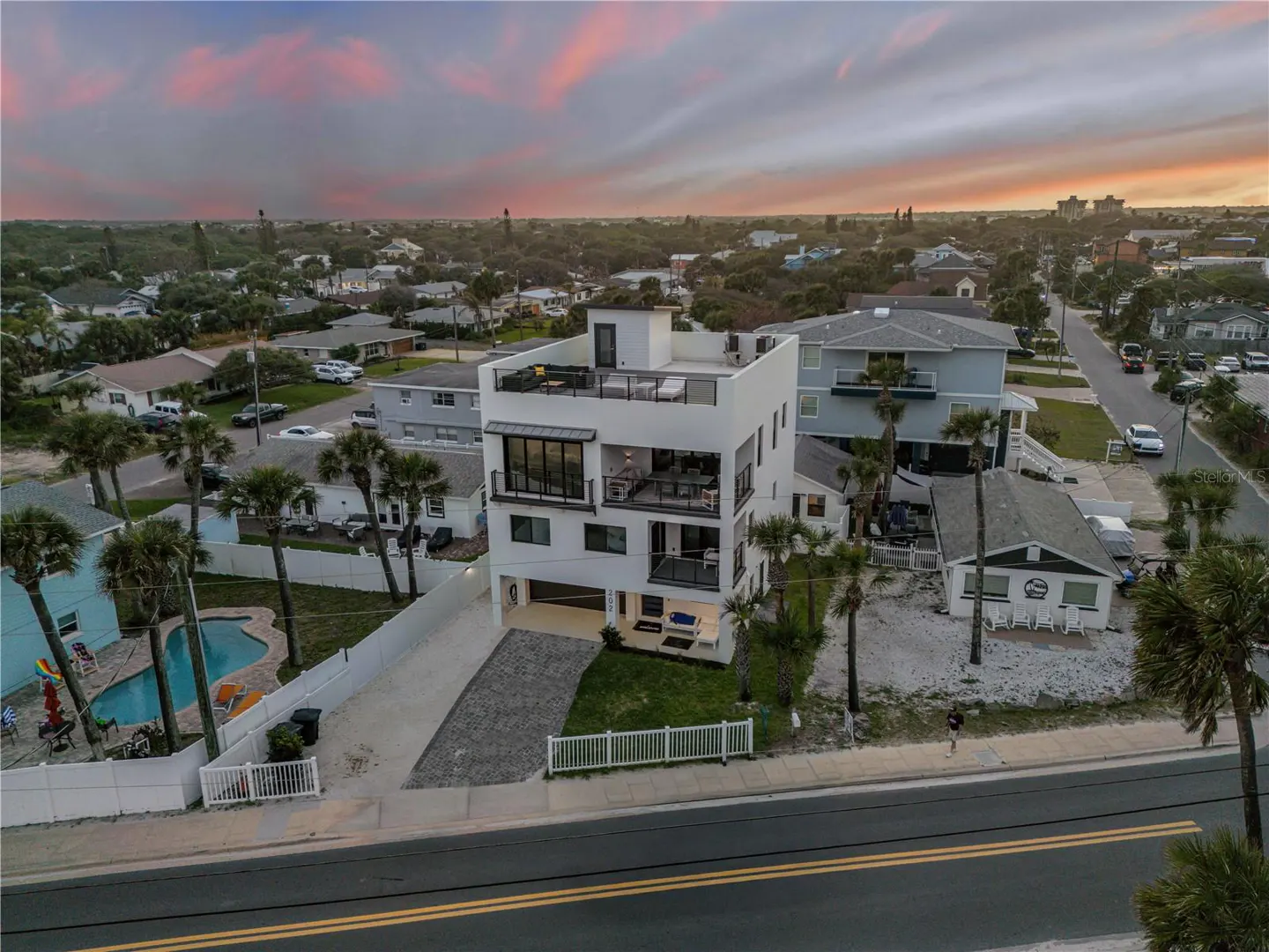 Aerial view of a modern, white, three-story beach house with a rooftop deck at sunset. Palm trees, pool, and other houses are visible.