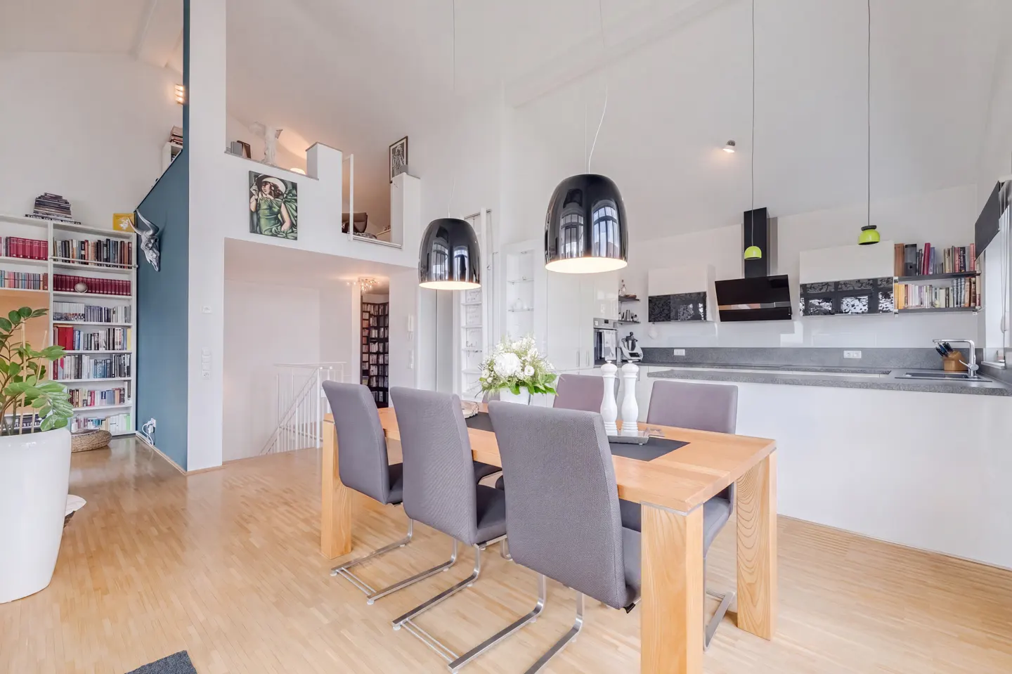 Bright, modern dining area with a wooden table, grey chairs, and black pendant lights. Open kitchen and bookshelves in the background.