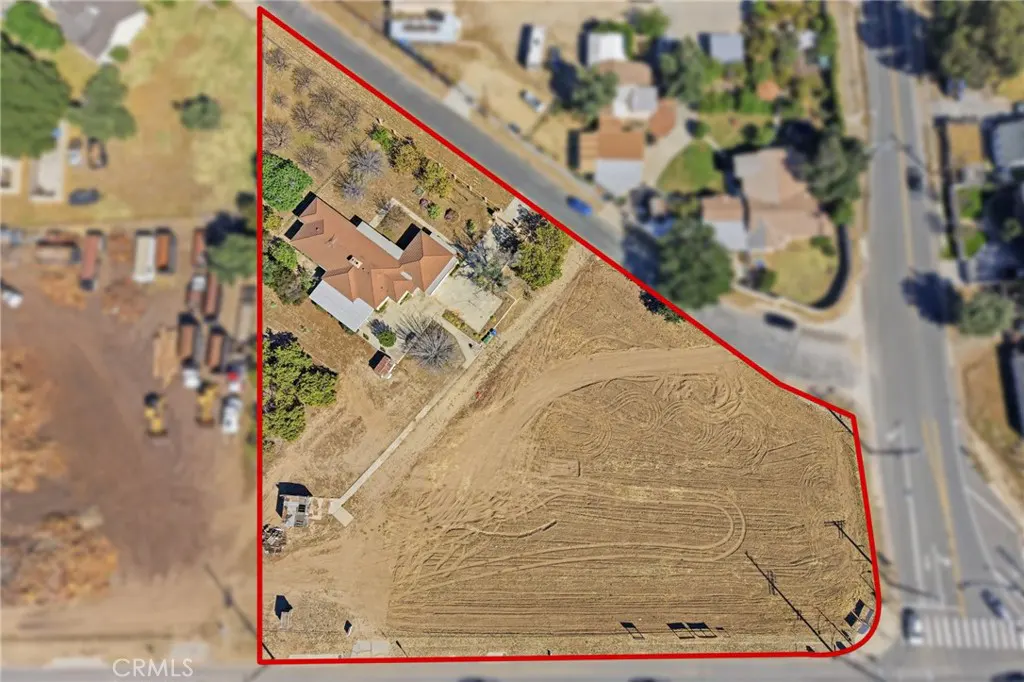 Aerial view of a large, vacant lot with a house, bordered in red. The lot is mostly dirt with tire tracks. A road runs along two sides.