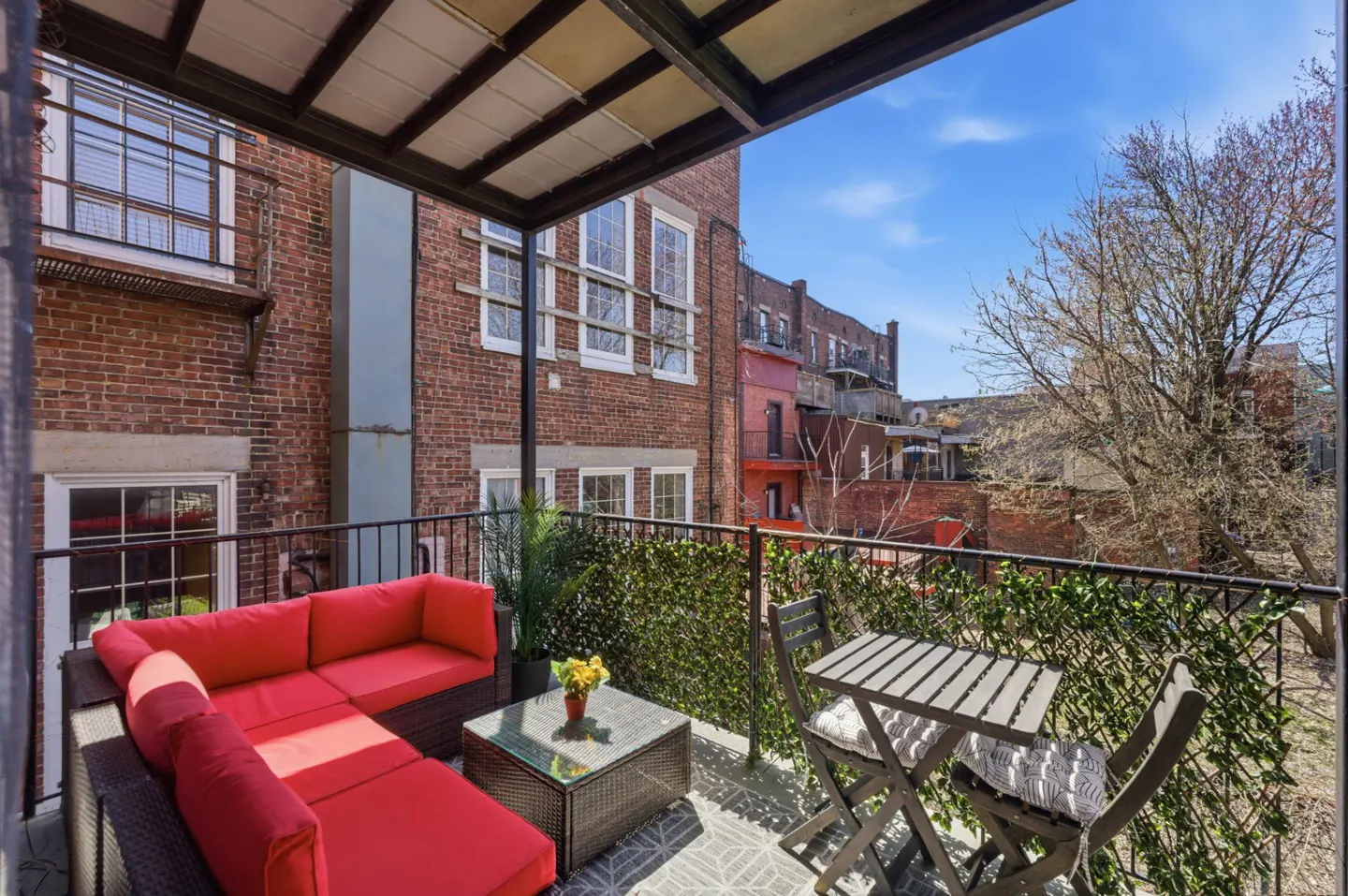 A balcony with a red couch, table, and chairs overlooks a brick building and a blue sky.