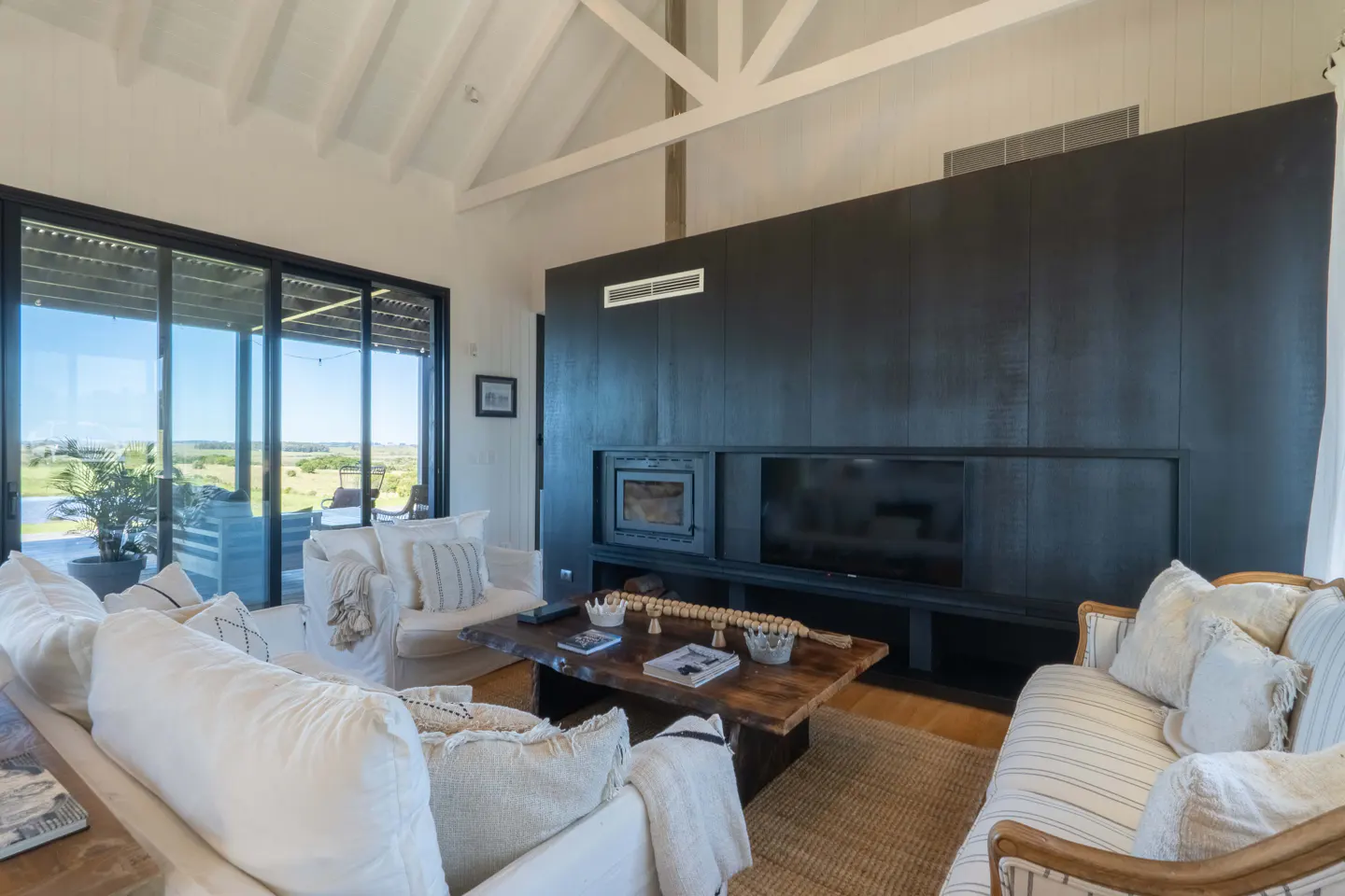 Bright living room with white sofas, wood coffee table, and black entertainment center with fireplace and TV. Large windows overlook a green landscape.