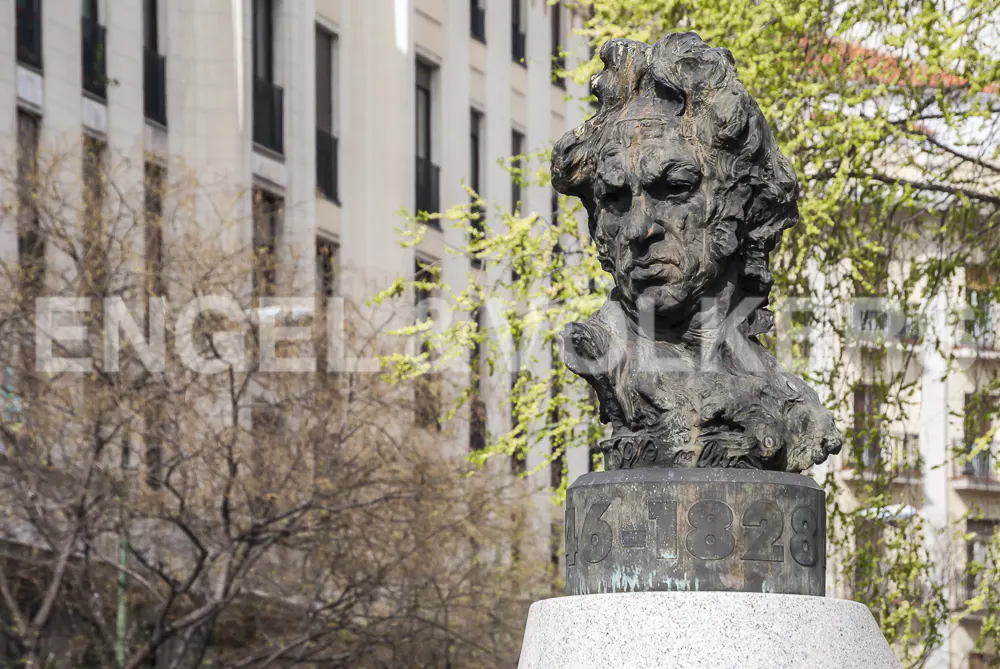 Bronze bust of Goya on a stone pedestal with "1746-1828" inscribed, set against a backdrop of buildings and trees.