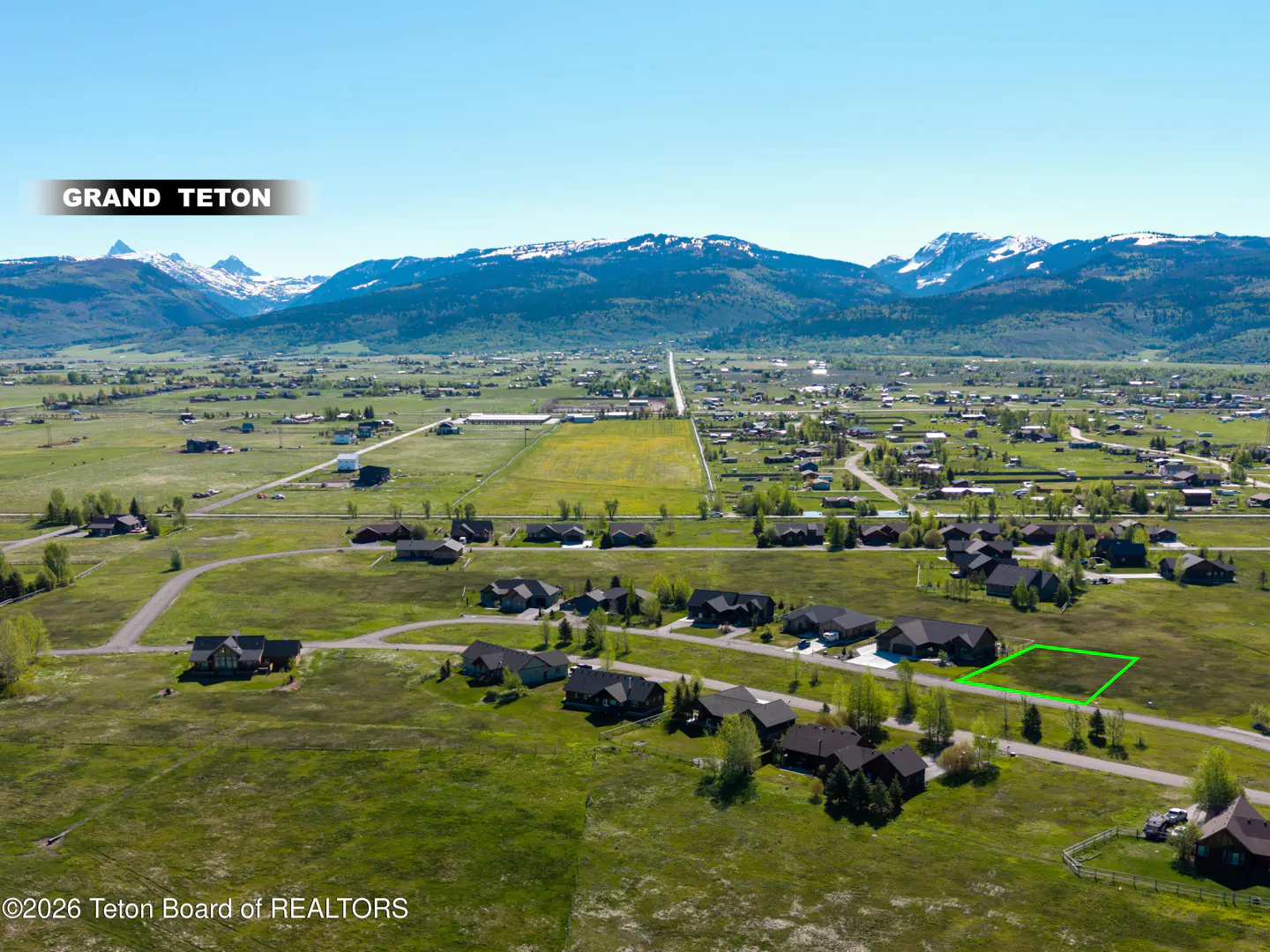 Aerial view of a green-bordered plot of land in a rural area with houses, fields, and the Grand Teton mountains in the background.