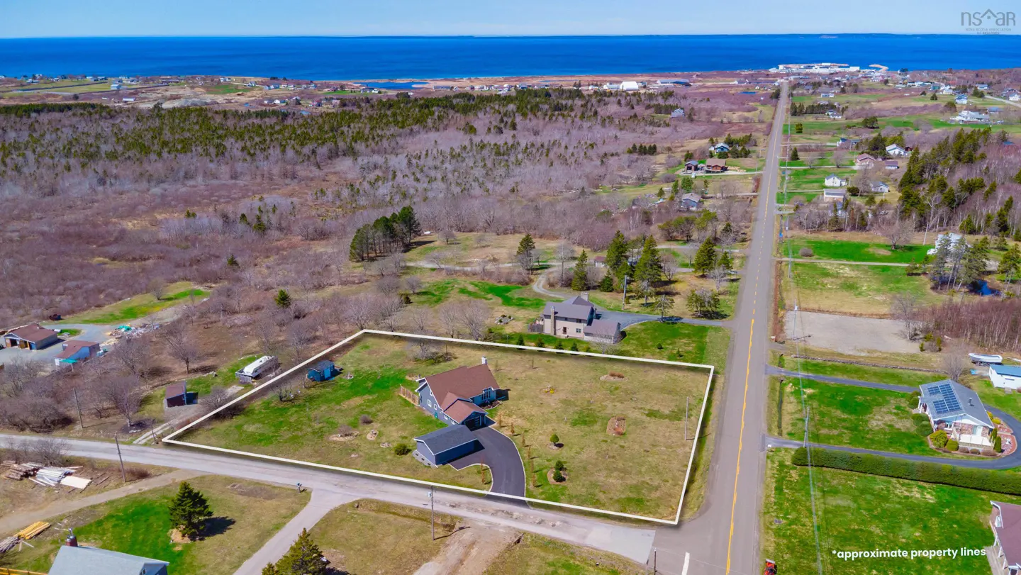 Aerial view of a property outlined in white, featuring a blue house with a brown roof, a driveway, and a green lawn. A road runs alongside the property, with trees and the ocean in the background.