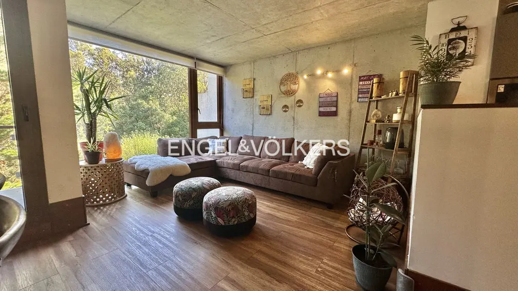 Living room with brown sectional sofa, floral ottomans, and wood floors. Large window with green trees outside. Concrete wall with decorations.