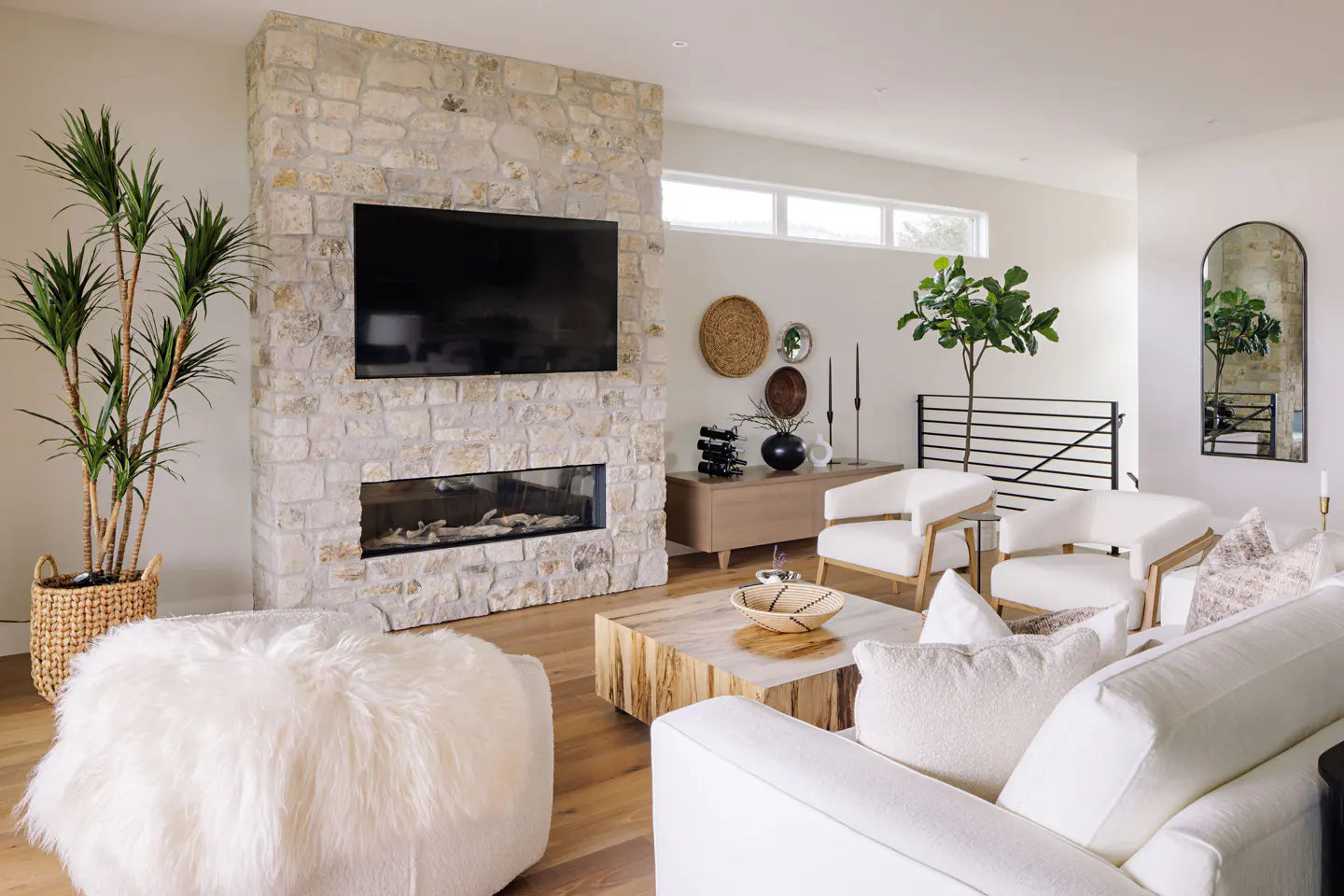 Bright living room with stone fireplace, TV, and modern furniture. White sofa, chairs, and a fluffy ottoman create a cozy atmosphere.