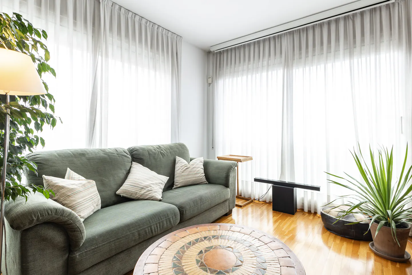 A bright living room with a green sofa, patterned round coffee table, potted plants, and floor-to-ceiling white curtains.