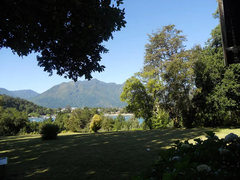 Scenic view of a lush green lawn with trees, mountains, and a lake in the background on a sunny day.