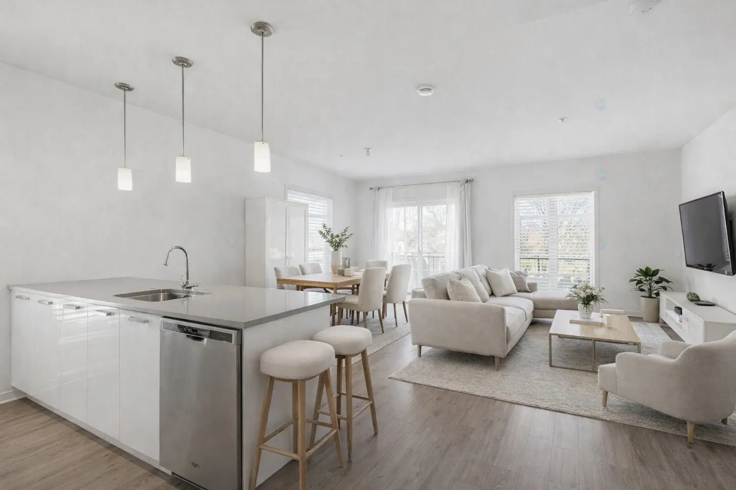 Bright, open-concept living space with white walls, wood floors, and modern furnishings. Kitchen island with stools, dining table, and living room with TV.