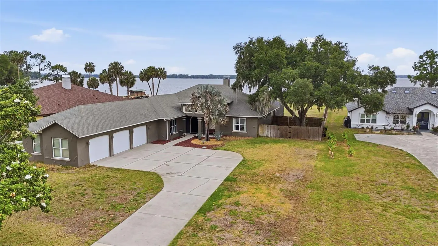 Aerial view of a gray single-story house with a three-car garage and a long driveway near a lake.