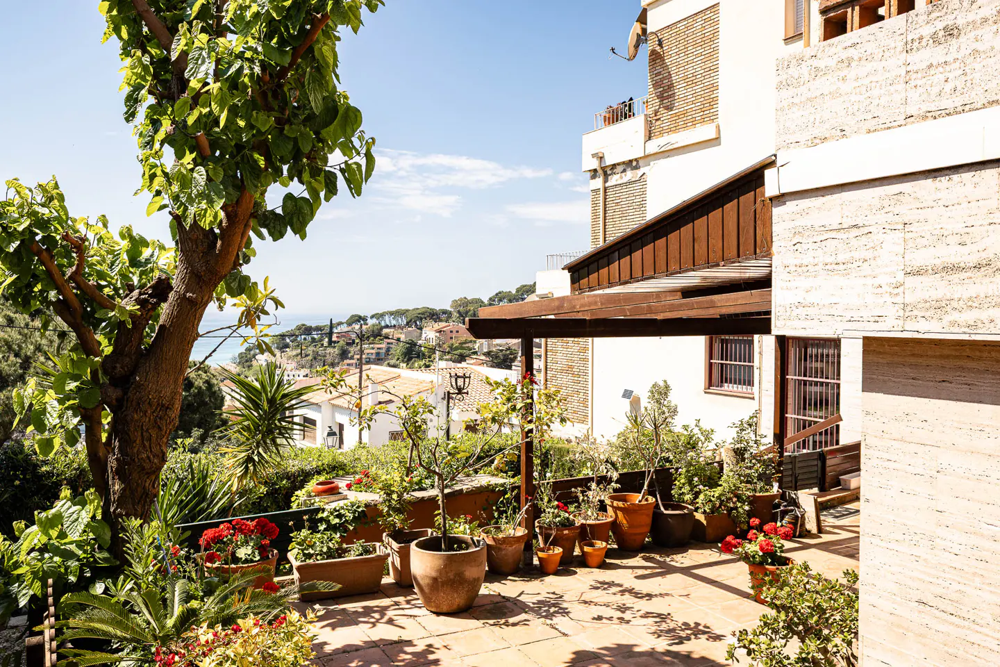 Sunny patio with potted plants and flowers, overlooking a town with red tile roofs and the ocean in the distance.