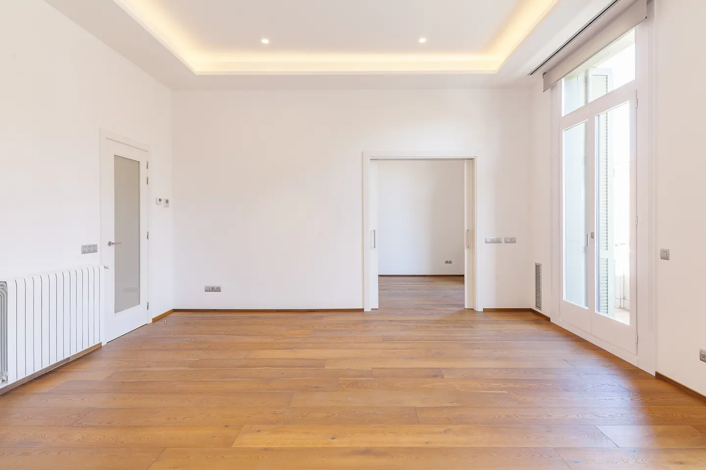 Empty room with white walls, warm wood flooring, recessed ceiling lighting, and a glass door leading to a balcony.