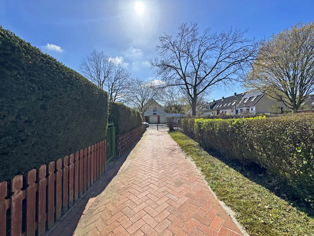 Brick path leads to a house, bordered by hedges and a fence under a sunny sky.