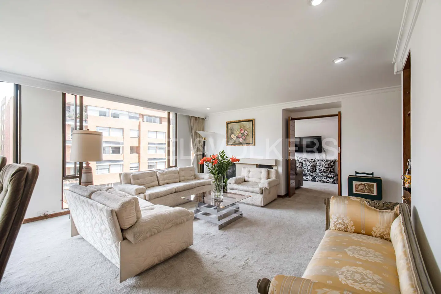 Bright living room with white sofas, glass table, and large windows overlooking a city building. Orange flowers add a pop of color.