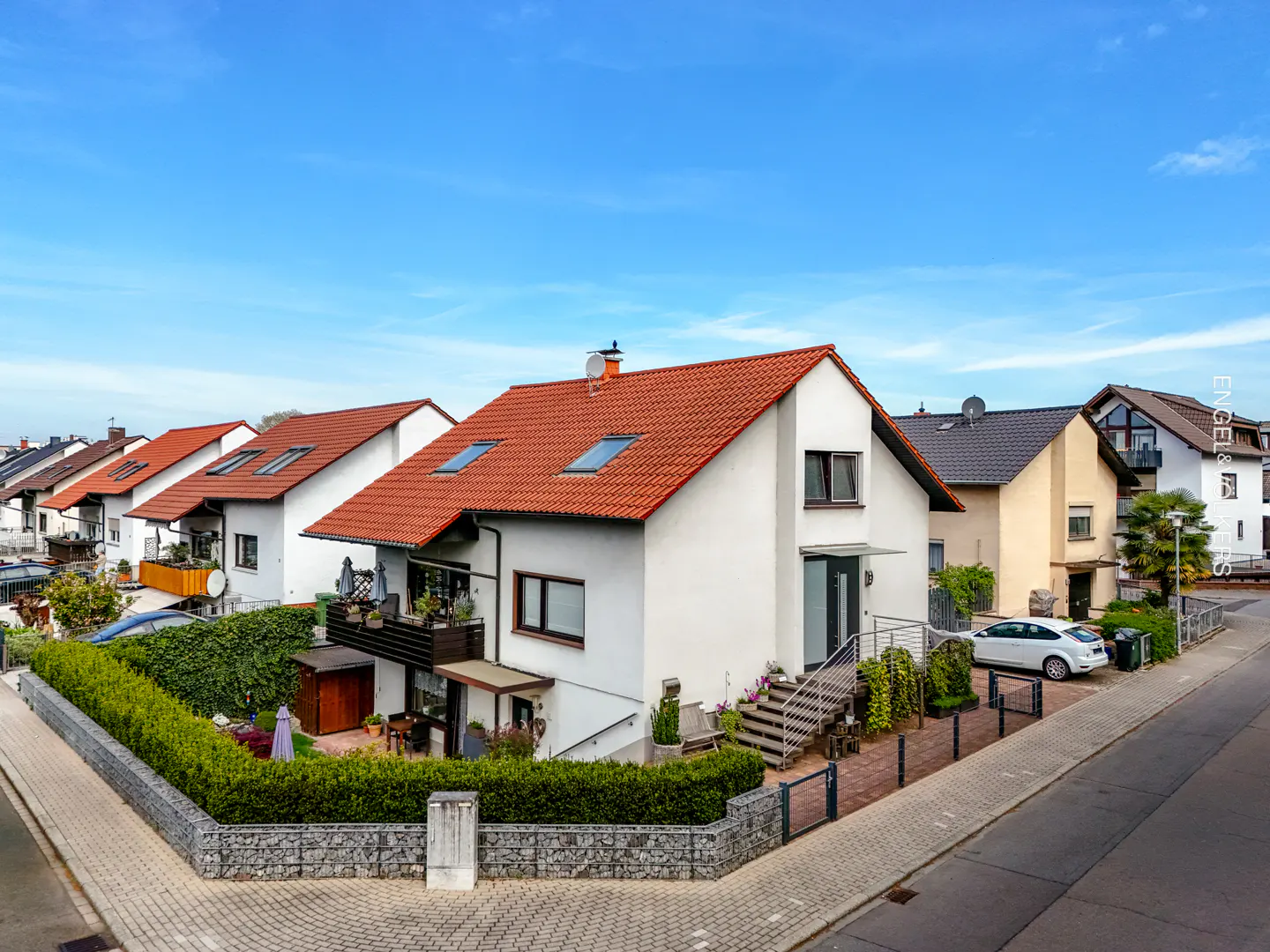 A white two-story house with a red tile roof, balcony, and a manicured hedge-lined yard on a sunny residential street.