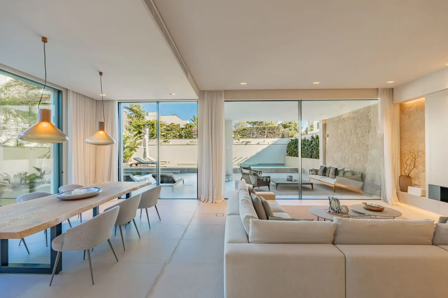 Bright, open-plan living and dining area with a large white sofa, wooden table, and floor-to-ceiling windows overlooking a pool and patio.