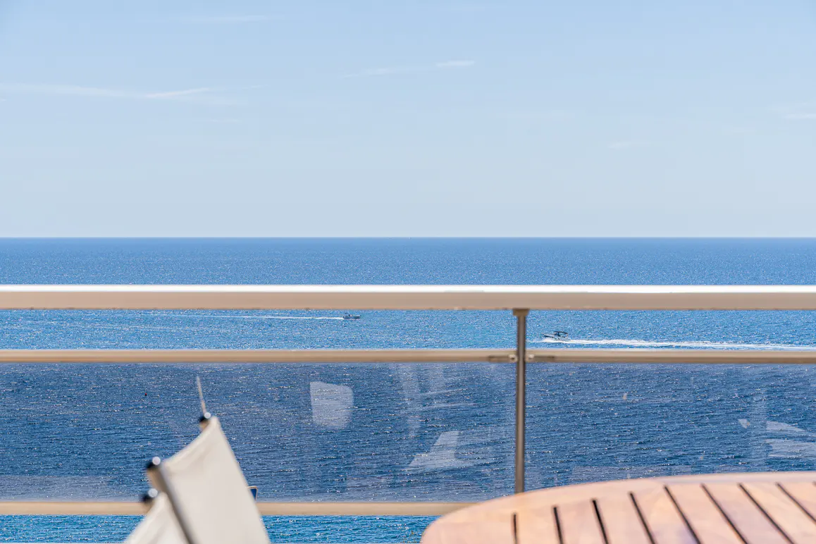 Ocean view from a balcony with a glass railing, a wooden table, and a chair. The sky is blue.