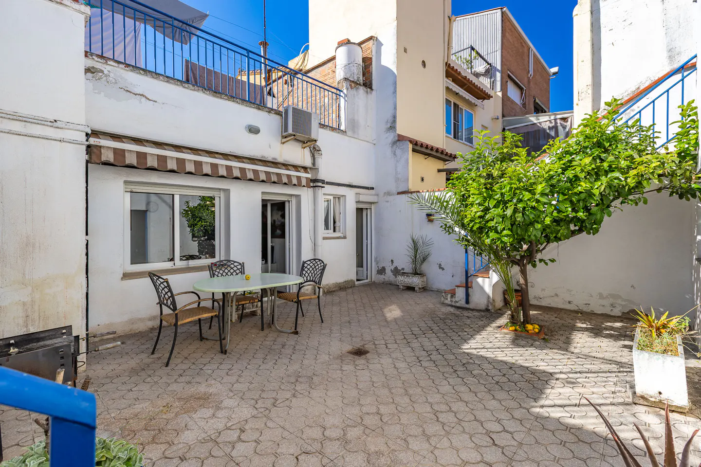 Outdoor patio with a table and chairs on a patterned stone surface, surrounded by white buildings and greenery.