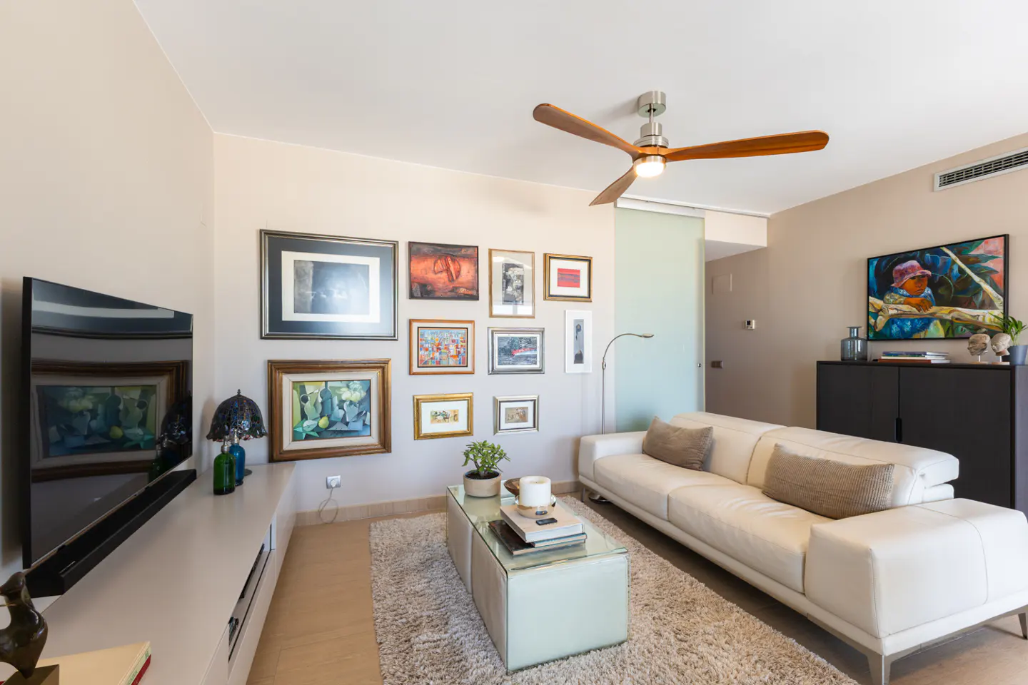 Living room with a white sofa, glass coffee table, and gallery wall with various framed artworks. A wooden ceiling fan is visible.