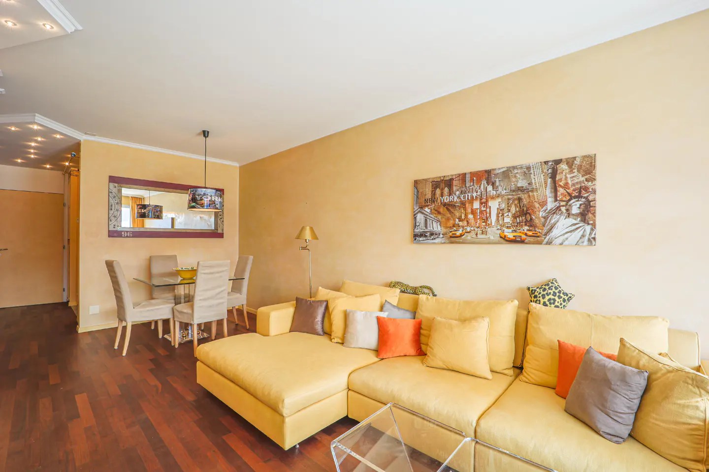Living room with yellow walls, a large yellow sectional sofa with pillows, and a glass dining table with chairs. A New York City painting hangs on the wall.
