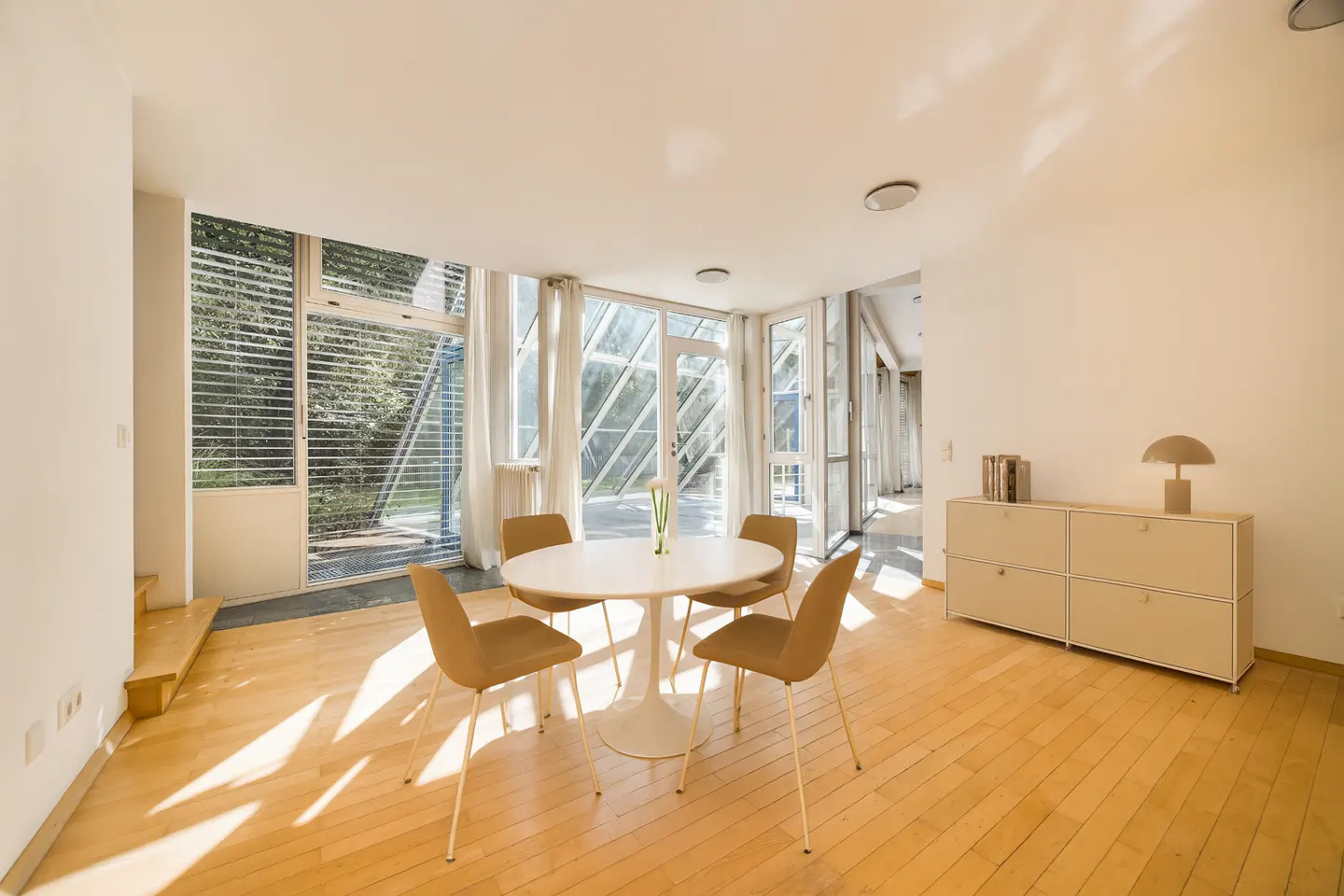 Bright, modern room with wood floors, a round table with four chairs, and floor-to-ceiling windows with a view of greenery.