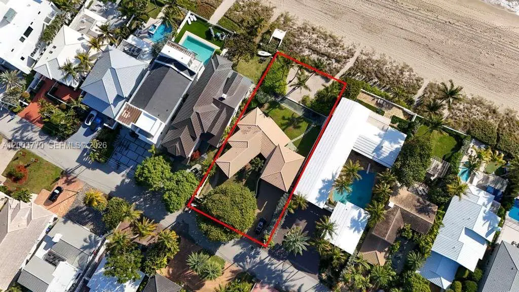 Aerial view of a tan-roofed house outlined in red, surrounded by lush greenery and neighboring homes near a sandy beach.