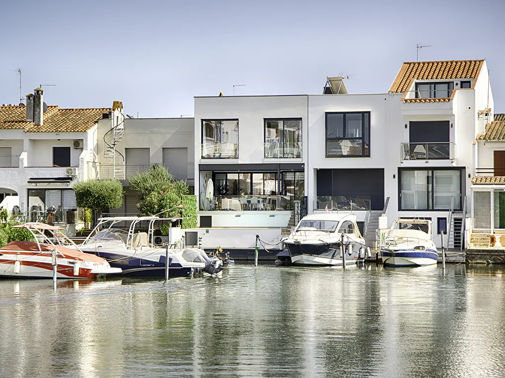 Waterfront homes with boats docked in front. White buildings with red tile roofs line the canal under a clear blue sky.