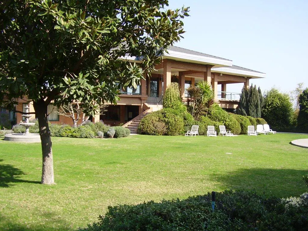 Exterior view of a tan house with a large green lawn, trees, and white lounge chairs.