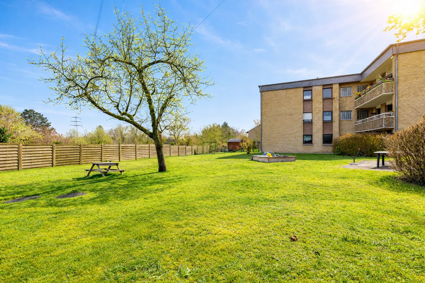 Exterior view of a yellow brick apartment building with a green lawn, picnic table, and tree.