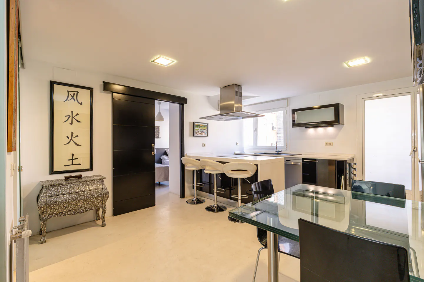 Modern kitchen with white island, three bar stools, black sliding door, and a glass dining table in a bright open space.