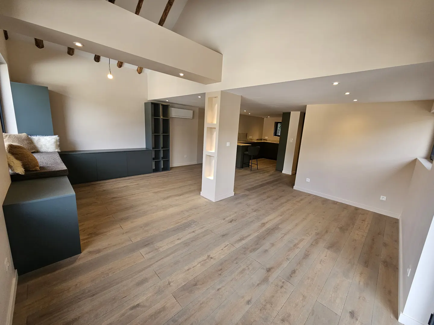 Open-concept living space with light wood floors, beige walls, and a high ceiling. A blue built-in bench and shelving unit are on the left.