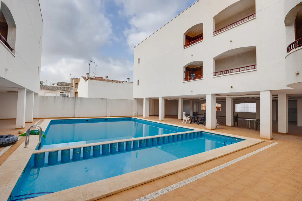 A rectangular blue swimming pool sits between two white buildings with balconies.