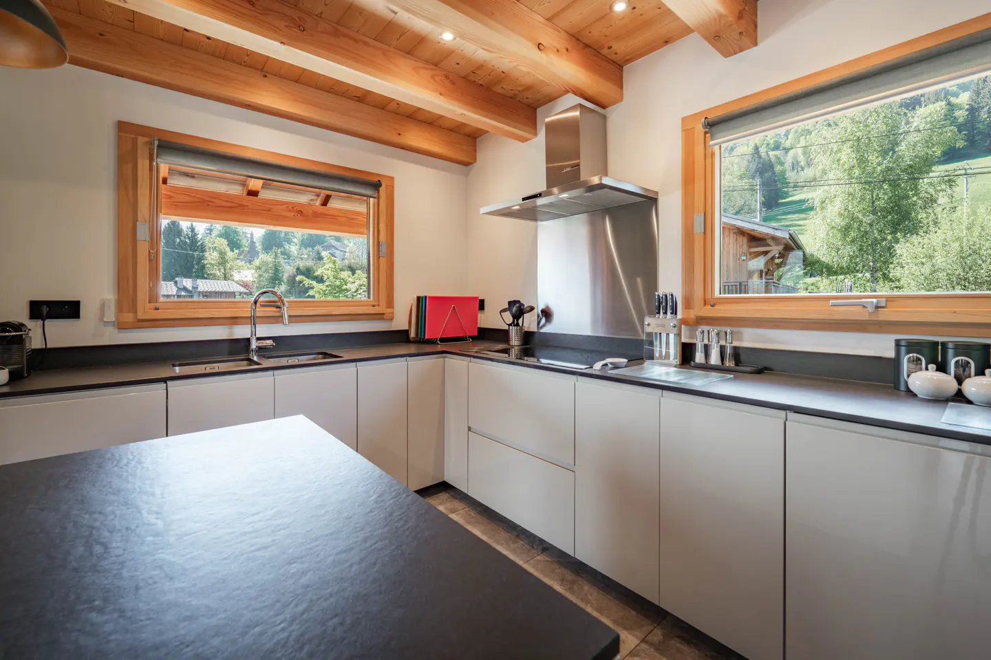 Bright kitchen with wooden beams, gray cabinets, and black countertops. Windows offer views of green trees and a house. Stainless steel range hood above the stove.