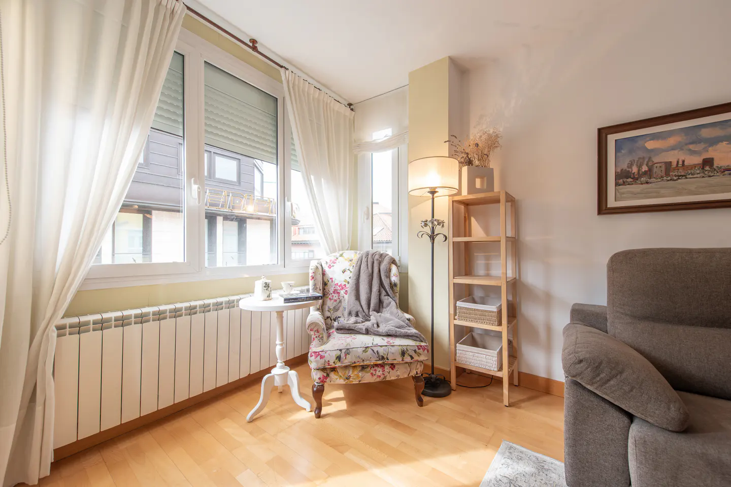 Bright living room with floral armchair, white curtains, and wooden floors. A small table and bookshelf add charm.