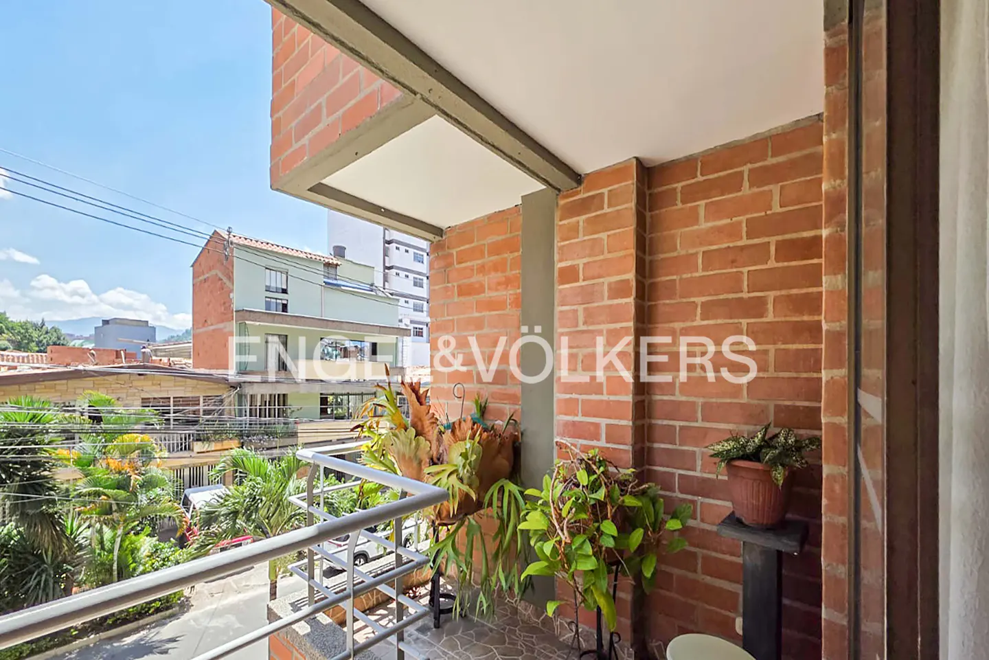 Balcony view with brick walls, plants, and Engel & Völkers logo. Cityscape visible in the background under a bright sky.
