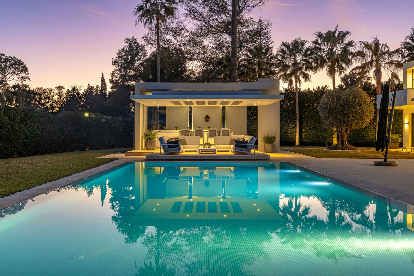 Outdoor pool reflecting a modern cabana with seating at dusk. Palm trees and lush greenery surround the property.