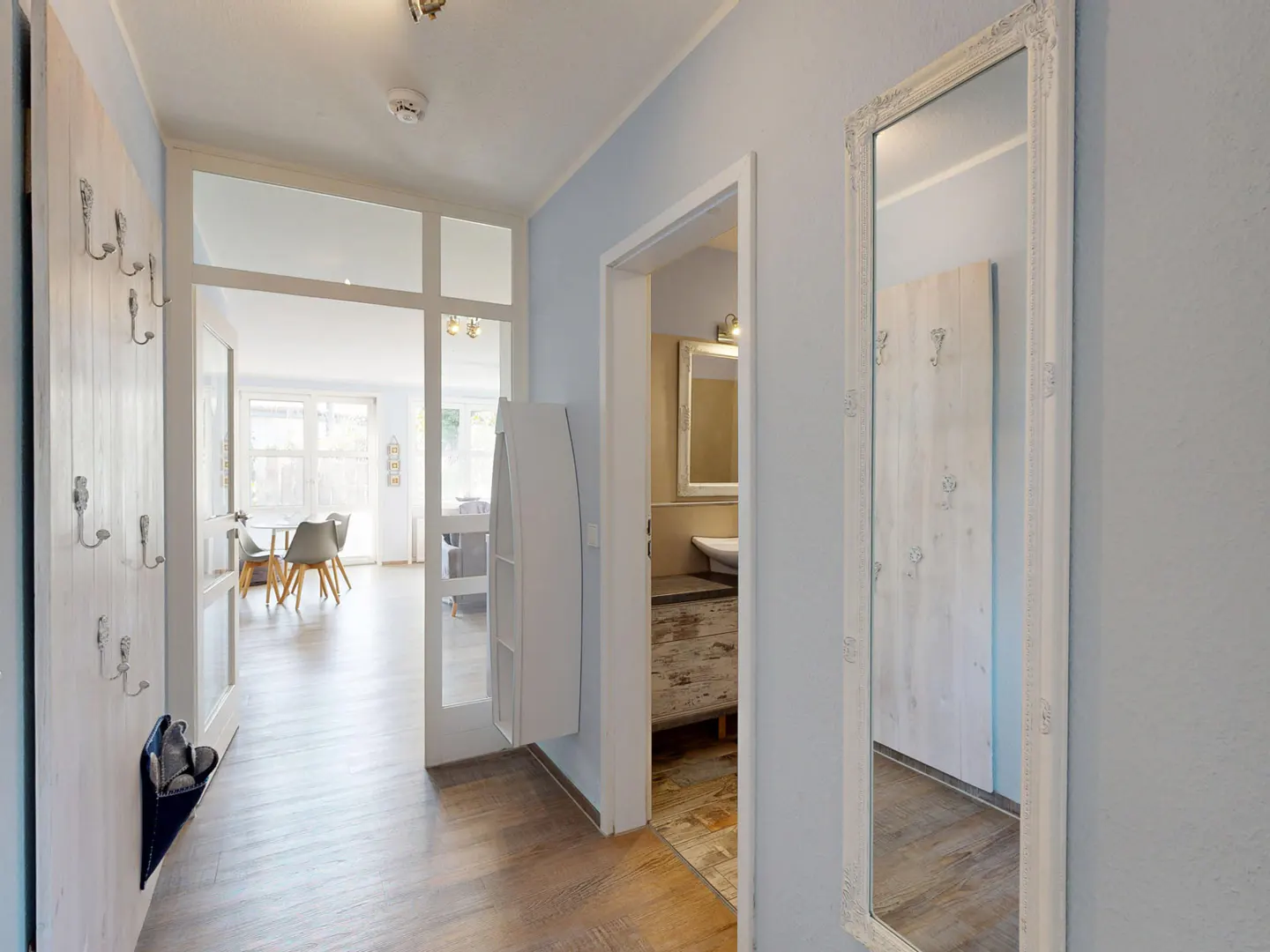 Hallway with light blue walls, wood floors, and white trim. A mirror hangs on the right wall. A doorway leads to a bathroom. A coat rack is on the left wall.