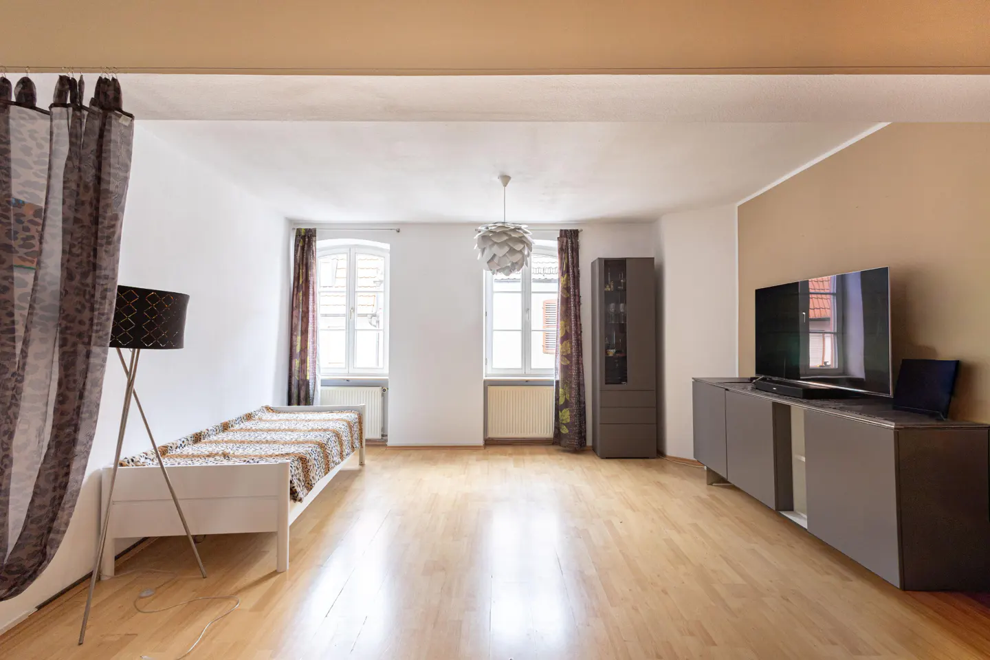 Bright living room with hardwood floors, white walls, and two windows. A daybed, floor lamp, TV, and cabinet furnish the space.