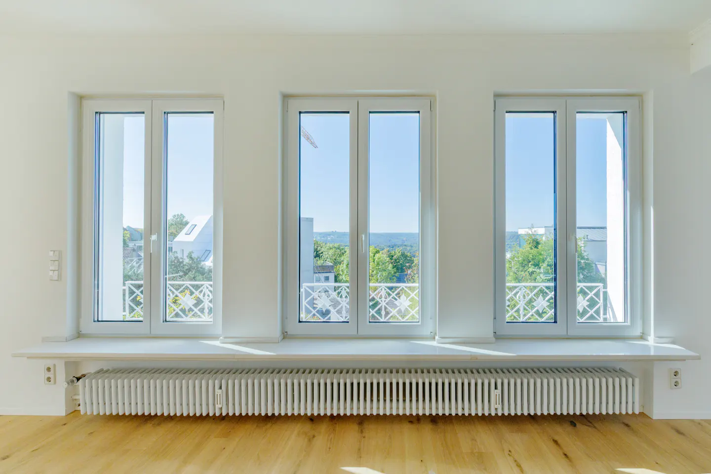 Three white-framed windows with a view of trees and buildings, above a white radiator on a wood floor.