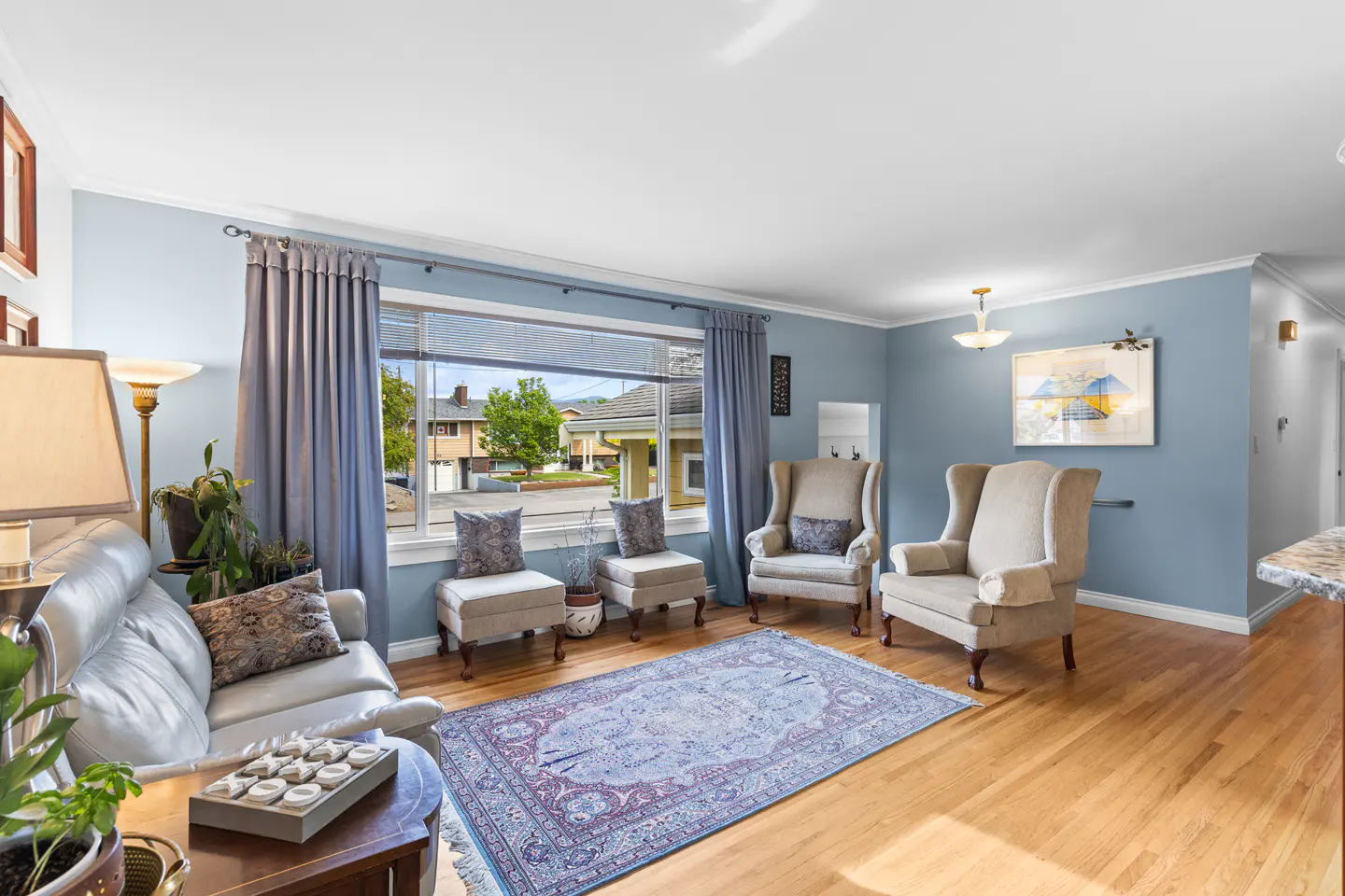 Living room with blue walls, hardwood floors, and a large window. Two armchairs and two ottomans sit on a blue patterned rug. A white sofa is on the left.