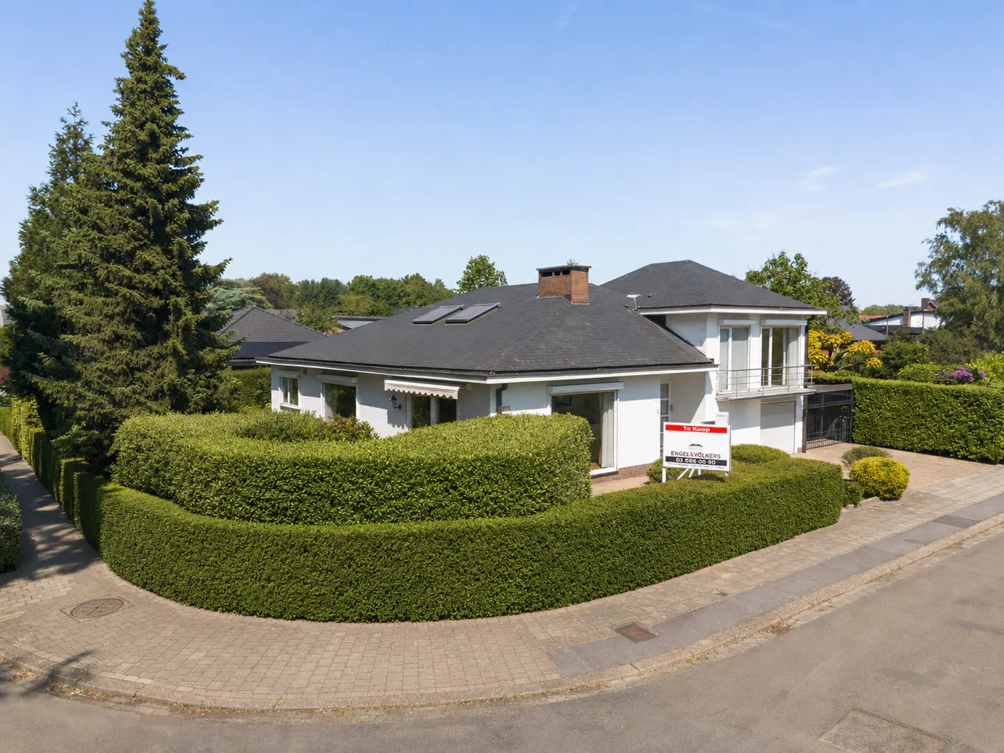 A white house with a dark roof is surrounded by a green hedge and trees. A "For Sale" sign is in front of the house.