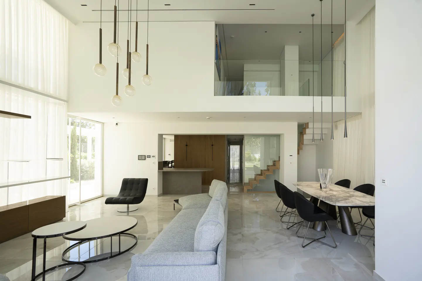 Bright, modern home interior with a gray sofa, marble table, black chairs, and a second-floor loft. Tall windows and pendant lights add to the airy feel.