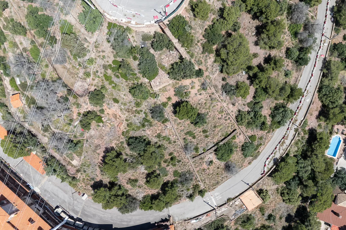 Aerial view of a hillside with trees, a road winding around it, and a blue swimming pool visible. Buildings with orange tile roofs are at the bottom.