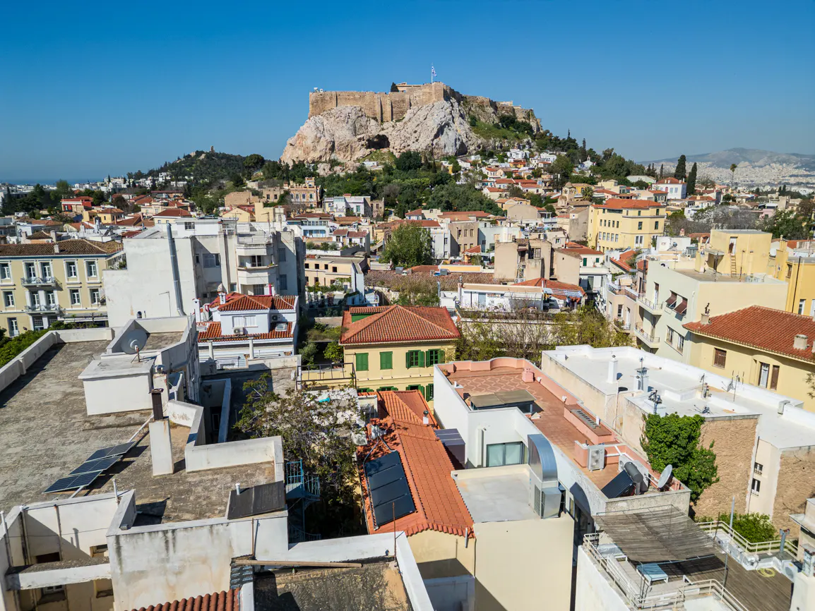 Athens cityscape under a clear blue sky, featuring rooftops and the Acropolis in the background.