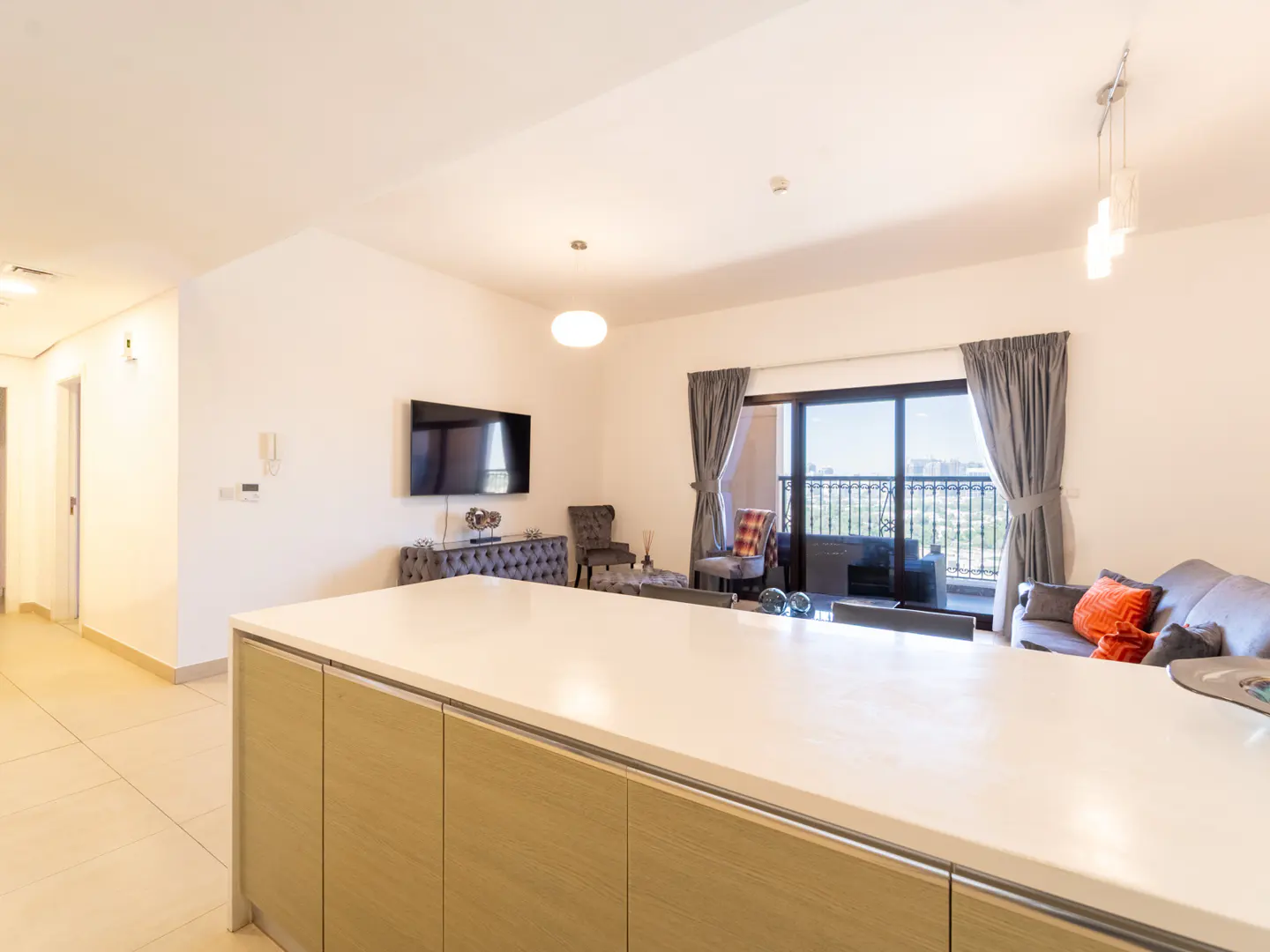 Modern open-plan living room with a white kitchen island, grey sofa, wall-mounted TV, and balcony with city views.