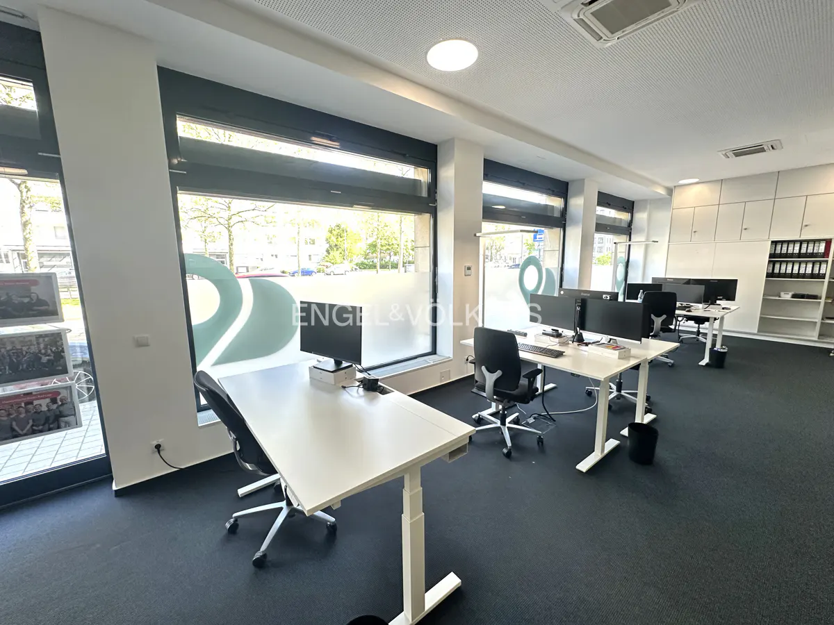 Bright office space with white desks, black chairs, and computers. Large windows with "Engel & Volkers" logo. Dark carpet and white storage cabinets.